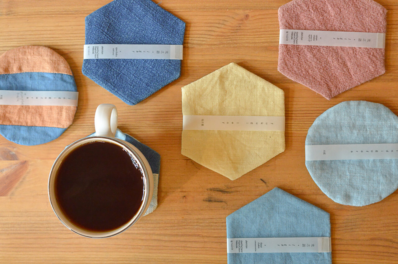 六角形や丸形のカラフルな布製コースターとコーヒーカップを木製テーブルに配置した様子